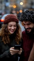 Young couple laughing while looking at smartphone together on city street with warm bokeh lights in background during evening hours.