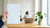 Hand holding blank white paper sheet in modern office workspace with blurred coworker and green plant, suitable for mockups, presentations, and corporate communications.