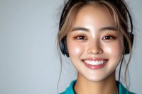 Young Asian woman wearing headphones smiles brightly against light background, showing natural glowing skin and genuine happiness in casual portrait.