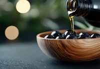 Fresh black olives in wooden bowl with pouring extra virgin olive oil, bokeh lights background creating luxurious food atmosphere.
