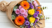 Colorful flower bouquet with orange gerberas, yellow roses, purple lisianthus and pink chrysanthemums wrapped in kraft paper on white wooden background.