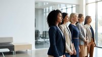 Diverse group of professional women standing in modern office corridor with glass partitions and natural light from windows, representing corporate teamwork and business leadership.