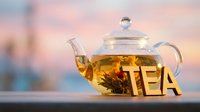 Glass teapot with blooming flower tea and wooden TEA letters on table against soft pastel sunset sky background.