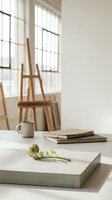 Minimalist workspace with wooden easel, stacked books, ceramic mug, and fresh green plant on white concrete table near bright window with natural light for creative projects.