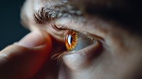 Extreme macro close up of amber colored human eye with detailed eyelashes and finger touching corner, showing intricate iris patterns on dark background.