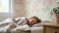 Young blonde woman peacefully sleeping in cozy bedroom with floral wallpaper and natural morning light streaming through window for wellness lifestyle content.