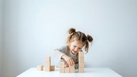 Young girl with pigtails playing with wooden building blocks at white table, developing fine motor skills and creativity through educational toy play.