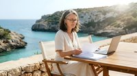 Middle-aged Caucasian businesswoman with glasses working on laptop at coastal terrace overlooking turquoise Mediterranean sea during golden hour vacation.