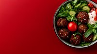Asian meatballs with teriyaki glaze served in black bowl with fresh vegetables, cherry tomatoes and herbs on vibrant red background. Copy space for menu.
