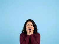 Young woman with long dark wavy hair and South Asian ethnicity wearing burgundy sweater, expressing surprise and excitement with open mouth and hands on face against blue background.