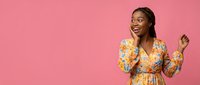 Young African American woman in colorful floral dress celebrating success with raised fists against pink background, expressing joy and achievement.