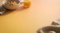 Overhead breakfast setup with cereal bowl, orange juice glass, and linen napkin on warm peachy background for food styling and morning meal content.