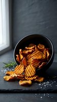 Crispy golden potato chips spilling from black bowl on dark slate surface with rosemary garnish and sea salt for gourmet snack presentation.