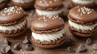 Chocolate macarons filled with swirled vanilla cream, decorated with salt flakes and scattered chocolate chips, photographed in rustic setting with soft lighting.