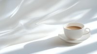 White porcelain cup filled with coffee or hot chocolate on saucer against soft white fabric background with dramatic diagonal shadows creating minimalist composition.