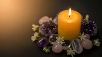 Burning orange candle surrounded by amethyst crystals, rose quartz stones and small flowers creating peaceful meditation and wellness atmosphere.