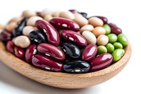Colorful variety of raw beans and legumes in wooden spoon, including kidney, black, navy and green beans, close up with selective focus on natural background.