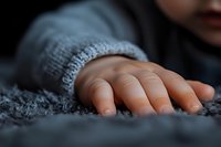 Close-up view of small child's hand in gray knitted sleeve touching soft textured fabric or carpet, creating intimate and tender mood in dark setting.