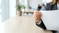 Businesswoman holding blank white paper document at wooden desk in modern office environment with colleagues working in background.