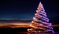 Glowing spiral light trail forming Christmas tree shape against dramatic sunset sky, creating magical holiday atmosphere with purple and orange illumination.