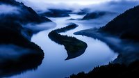 Mystical aerial view of winding river through mountain valley at dawn with low-lying fog creating ethereal atmosphere in moody blue tones. Dramatic landscape photography.