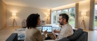 Young African American woman and Caucasian man working together on laptop in modern living room with warm lighting and large windows for remote work.