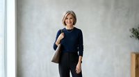 Mature blonde woman in professional business casual attire with navy sweater and black pants, carrying brown shoulder bag, standing confidently in modern office environment.