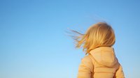 Young woman with blonde hair in yellow winter puffer jacket against bright blue sky, wind blowing through hair, back view, capturing freedom and adventure.