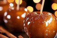 Glossy caramel apple with wooden stick on dark background with golden bokeh lights, close up view showing sweet candy coating texture and reflections.