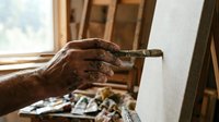 Artist hand holding paintbrush creating artwork on canvas in sunlit studio workspace for creative projects and artistic expression.