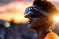 Young African American woman wearing virtual reality headset against dramatic sunset sky, orange glow illuminating her profile as she explores digital world.