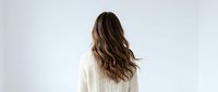 Young woman with long wavy brown hair wearing white knit sweater standing with back turned against neutral background for lifestyle content.