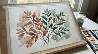 Watercolor botanical illustration featuring autumn leaves in warm orange and cool green tones on textured paper, displayed in wooden frame on artist workspace.