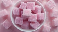 Pink sugar cubes coated in fine crystals arranged in white ceramic bowl, top view macro shot with soft focus background creating dreamy dessert composition.