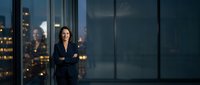Professional businesswoman with crossed arms standing confidently in modern office with city skyline view through large windows at evening.