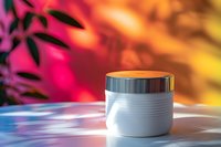 White cosmetic cream jar with metallic lid on reflective surface against vibrant pink and orange bokeh background with leaf shadows creates luxurious beauty product scene.