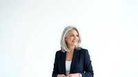 Mature woman with long gray hair wearing dark blazer and white top, smiling while looking away against white background. Suitable for corporate, business, and professional website use.