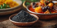 Ground black charcoal powder in rustic clay bowl on wooden table with blurred traditional dishes in background, natural food supplement and detox.