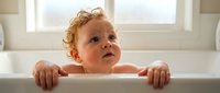 Adorable curly-haired toddler with blue eyes sitting in white bathtub during bath time, looking up with curious expression in bright bathroom setting.