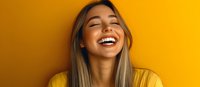 Young Asian woman with bright genuine smile and long blonde hair laughing with eyes closed against vibrant yellow background, expressing pure joy and happiness.