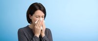 Woman with dark hair and fair complexion blowing nose into tissue against blue background, depicting cold, flu, or allergy symptoms for healthcare and wellness content.