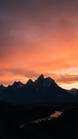 Dramatic mountain peaks silhouetted against vibrant orange and purple sunset sky with winding river valley below creating stunning landscape scenery.