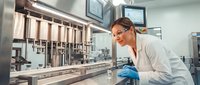 Female scientist in protective eyewear examining pharmaceutical manufacturing equipment in modern laboratory facility for quality control testing.