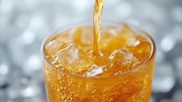 Refreshing cold drink being poured into glass with ice cubes, golden liquid splashing in transparent tumbler against blurred background, close up view.