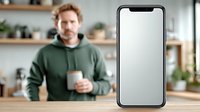 Young Caucasian man in green hoodie holding coffee cup in modern kitchen with blank smartphone screen for app mockup and digital marketing content.
