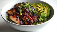 Glazed teriyaki chicken bowl with fresh sliced avocado, green onions and black sesame seeds served on mixed greens in white ceramic bowl.