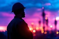 Silhouette of bearded construction worker in hard hat against vibrant purple and blue sunset sky with blurred city lights creating dreamy urban backdrop.
