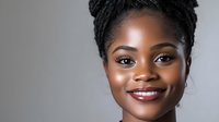 Young African American woman with natural braided hairstyle and radiant smile against gray background, showcasing confident and professional appearance for beauty and lifestyle content.