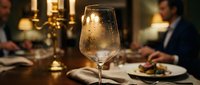 Elegant wine glass on restaurant table with warm candlelight and blurred diners in background creating sophisticated dining atmosphere for hospitality marketing.