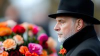Senior Caucasian man in black hat and coat with gray beard in profile view next to vibrant bouquet of roses and peonies at formal event or celebration.